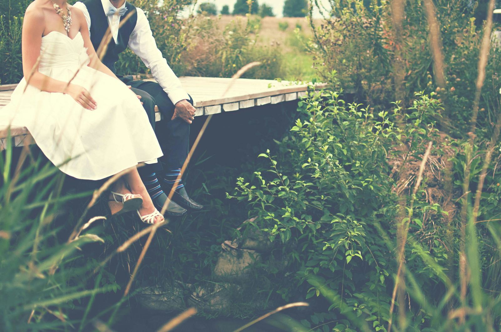 A bride and groom sitting side by side at the end of a timber jetty, framed by long grass
